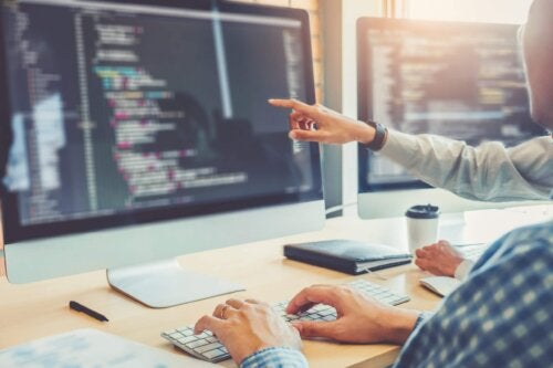 Image showing two coders looking at monitor with code on the screen. One of the coders is pointing at the monitor, the other has hands on keyboard.