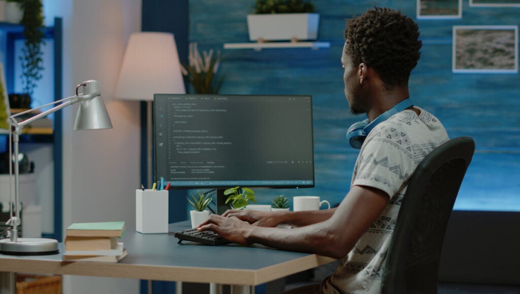 Young black developer with blue earphones resting around neck on shoulders sitting in home office at desk with hands on keyboard and monitor displaying code.