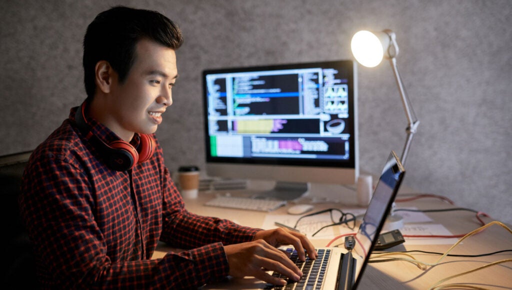 Desarrollador sonriente escribiendo en una computadora portátil con resultados de pruebas automatizados que se muestran en un monitor dual a su izquierda.