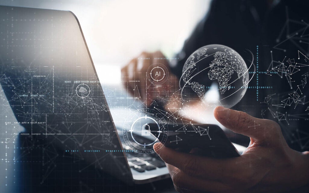 Image of a person’s hands holding a cellphone near a laptop with a digital overlay of a lock, cloud, and globe.